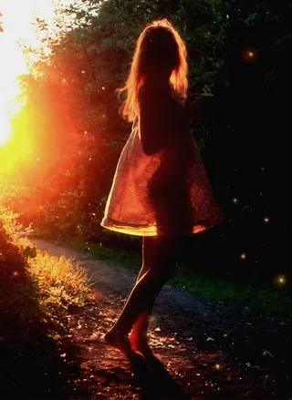 Silueta de una niña descalza con cabello largo vistiendo un vestido, de pie al aire libre al atardecer con contraluz y partículas de luz.