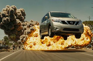 A photorealistic cinematic scene showing a Nissan Quest minivan airborne after jumping off a ramp, with a massive explosion and thick smoke erupting in the background on a city street.