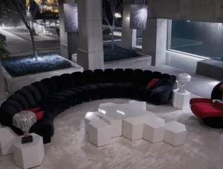 Modern living room featuring a black leather curved sofa with red cushions, white geometric modern tables, metallic tree decor, concrete walls, and a blue floor at night.
