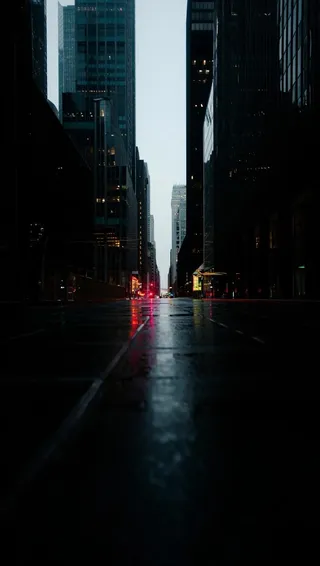 Empty city street at night with reflections of lights on wet pavement, tall skyscrapers lining both sides.