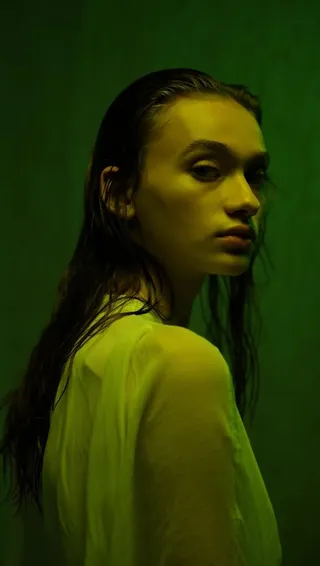 Portrait of a young woman with wet hair and a yellow tint on her skin, lit with moody green lighting and wearing a sheer top.