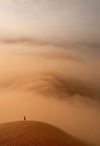 A lone figure standing on a desert dune enveloped in a vast, ethereal caramel-colored fog with soft shadows.