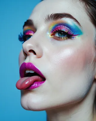 Close-up portrait of a woman with pale skin wearing a blue beret, vibrant rainbow glitter eyeshadow, magenta glossy lipstick, and playfully sticking her tongue out.