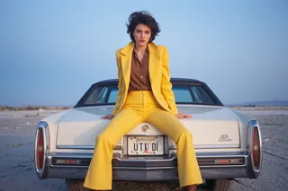 Kobieta w żółtej marynarce i dzwonowatych spodniach siedząca na bagażniku białego samochodu Cadillac z tablicą rejestracyjną Utah podczas niebieskiej godziny na solniskach, wietrzna pogoda.