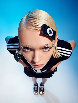 Close-up portrait of a blonde woman wearing a black Adidas outfit with white stripes, star earrings, and a retro iPod clipped to her sleek hair against a blue gradient background.