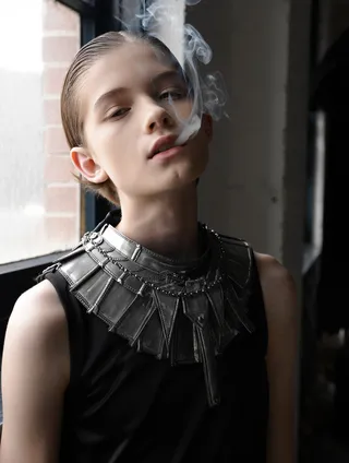 Portrait of a young person with short hair wearing a black sleeveless top and an intricate metal collar, exhaling smoke near a window.