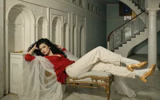 A 1950s female popstar with goth style reclining on a gold-accented bench in a marble-floored sophisticated interior with an ornate staircase in the background.