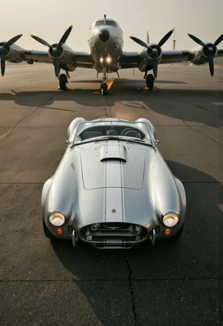 A silver vintage Cobra sports car with headlights on parked on an airport tarmac facing a large silver B-25 Mitchell bomber aircraft with four propellers under an overcast sky.