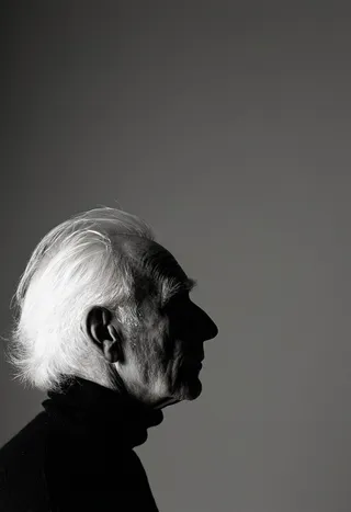 Dramatic black and white profile portrait of an elderly man with white hair wearing a black turtleneck, against a plain gradient background with side lighting.