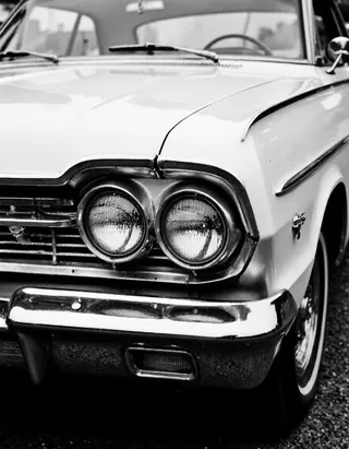 Black and white close-up photo showing the front left side of a classic 1960s car with prominent round headlights and chrome bumper.