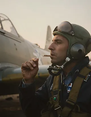 A tired pilot wearing vintage flight gear smoking a cigarette next to his fighter plane at early sunrise with cinematic and moody lighting.