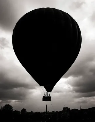 Black and white high-contrast photograph of a colossal hot air balloon silhouetted against a stormy sky over Paris, showcasing detailed basket and dramatic lighting.
