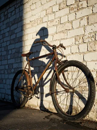 A rusted bicycle leaning against a weathered brick wall with its shadow showing a silhouette of a rider pedaling intensely, captured in warm late afternoon light.