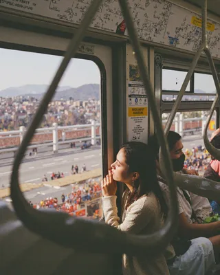 A woman sitting inside a subway carriage looking thoughtfully out the window, framed by a blurry foreground handle, with wiring diagrams and signs visible inside the carriage.