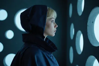 Side profile of a blonde woman wearing a navy blue cloak and hood, illuminated by white light from large circular patterns on a dark blue wall in dim lighting.