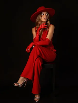 A confident woman in a vibrant red sleeveless jumpsuit, long red gloves, and a wide-brimmed red hat sitting on a black chair with silver high-heeled sandals on a dark background.