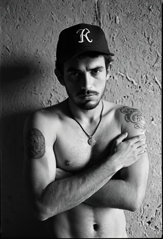 Black and white analog photo of a shirtless young man wearing a backwards baseball cap, showing tattoos on his arms and torso, standing against a rough textured wall with shadows.