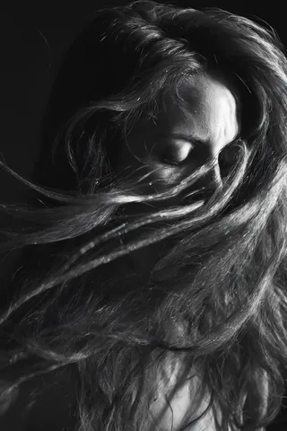 Black and white close-up portrait of a woman with long flowing hair covering most of her face, eyes closed, captured in dramatic motion with fine art lighting and high contrast.