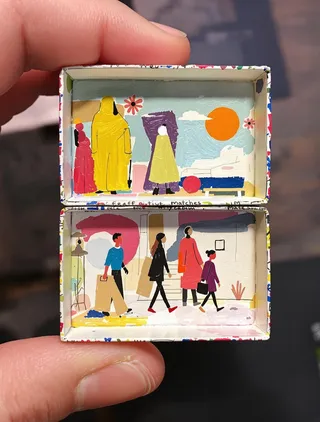 Hand holding a small matchbox painted inside with a modern art scene featuring minimalist sculptures and flat figures in vibrant candy colors with thick black outlines.