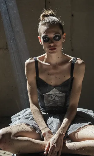 A candid portrait of a ballerina sitting cross-legged with dark makeup around her eyes, wearing a grey ballet dress covered in dust, illuminated by a light shaft highlighting dust motes.