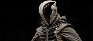 Close-up of a futuristic white helmet with a black visor and red line, paired with a gray cape featuring black metal panels, set against a black background.