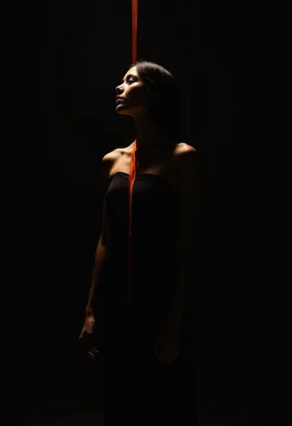 A solitary woman stands in deep shadow with a vibrant orange stripe bisecting her against a midnight black background, softly lit to highlight her profile and delicate posture.