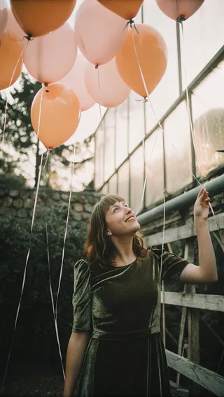 Young woman wearing a green velvet dress holds orange and pink balloons outdoors, smiling and looking up in soft natural light.