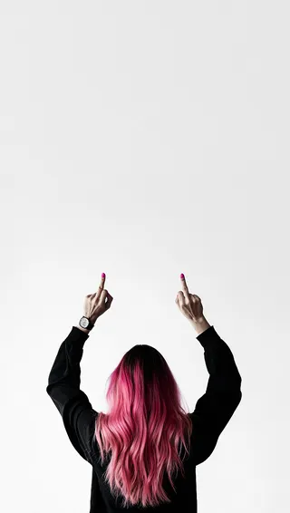 Person with long wavy pink hair wearing a black long-sleeve shirt, shown from behind with both middle fingers raised against a plain light background.