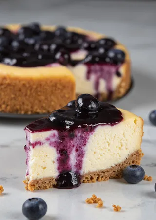 A close-up of a slice of blueberry cheesecake with a graham cracker crust, topped with glossy blueberry sauce and fresh blueberries, set on a neutral marble surface with soft natural lighting.