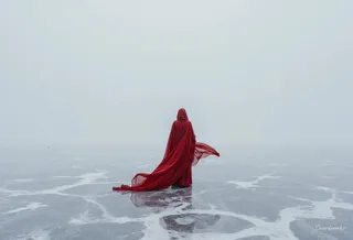 A solitary figure wearing a flowing red cape stands on cracked frozen ice beneath an overcast sky in a surreal, ethereal landscape.