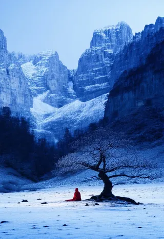 A lone figure in a crimson robe sits cross-legged beneath a gnarled tree in a snow-covered valley, with jagged indigo mountains rising in the background under twilight light.