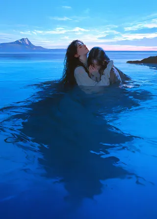 Two young adults with dark hair floating peacefully in a calm turquoise ocean under a blue sky with distant mountains.