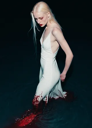 An ethereal albino woman with pale skin and white hair wading through a dark crimson liquid, wearing a backless silk white gown that is stained red from the hem, set against a dark background.