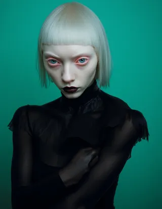 Eerie portrait of a young woman with platinum blonde bob hairstyle and blunt fringe, wearing a black sheer turtleneck, set against a vibrant teal background with light blue eyes and dark lip makeup.