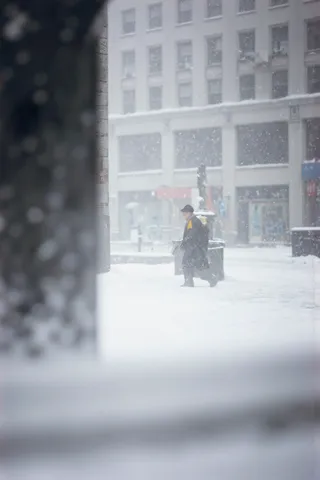 A silhouette of a man walking on a snow-covered city street during heavy snowfall with a bright yellow scarf, framed in a painterly, minimalist style inspired by Saul Leiter.