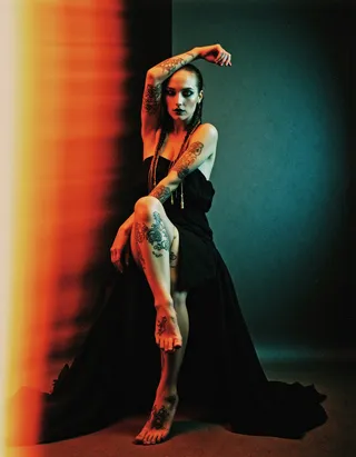 Full-body portrait of a pale-skinned woman with box braids decorated with beads, wearing a black maxi dress, showing gothic tattoos on her arms and legs, seated with arm raised against an abstract colorful background.