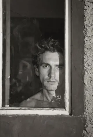 Black and white high contrast photograph of a man with messy short hair looking out a vintage window with part of his face in shadow, taken on Ilford Delta 400 film.
