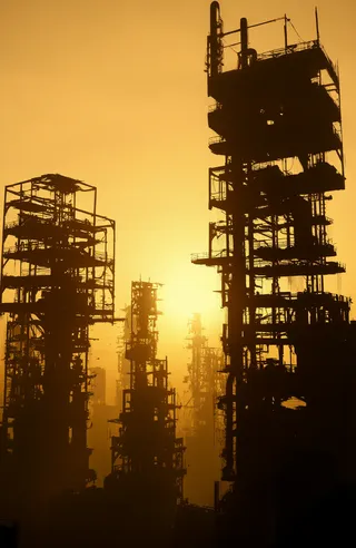 Silhouetted industrial cityscape with tall skeletal buildings and metal frameworks against a warm golden-orange sunset sky with hazy mist effects.