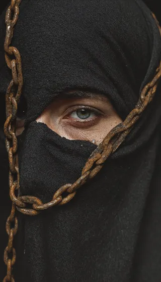Close-up portrait of a woman with lost identity theme, her face mostly covered by black cloth, an antique rusted iron shackle draped across, highlighting textured rust and one visible eye.