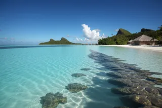 Clear turquoise lagoon in Bora Bora with overwater bungalows, palm trees casting shadows on coral reef under blue sky with white clouds.
