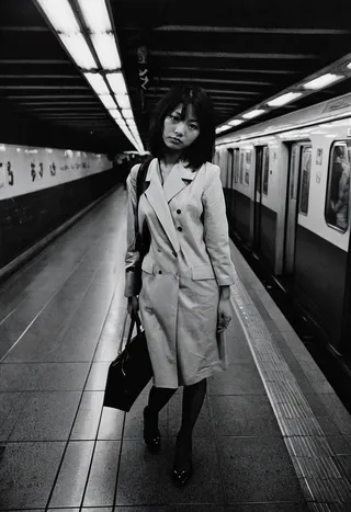 A black and white photo of a woman standing on a subway platform in Tokyo, wearing a long coat and carrying a handbag, with a train on the right and fluorescent lights above.