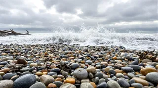 Close-up of a shoreline covered in smooth, multicolored pebbles with foamy waves crashing under an overcast sky with scattered driftwood and seaweed