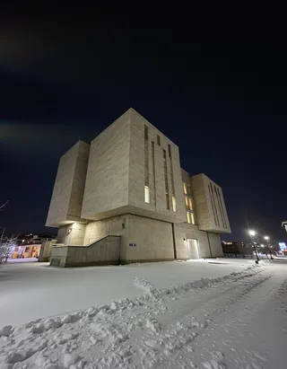 Brutalist concrete opera house illuminated with warm lighting at night, with snow-covered ground in the foreground under a dark sky.