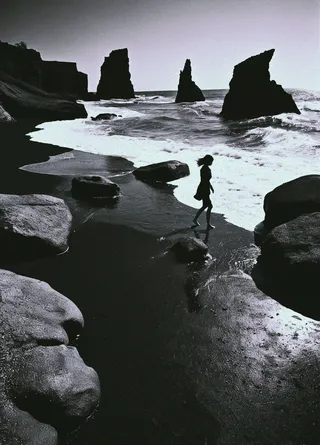 Silhouette of a woman walking on a black sand beach with rocky formations and waves breaking in monochrome style, spotlight highlighting her figure.