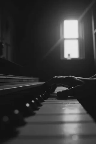 Black and white low angle close-up of a person's left hand playing piano keys in a dimly lit room with a blurred window in the background