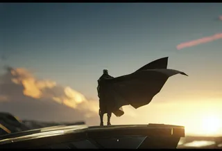 Distant view of a faceless male in a futuristic suit with a long flowing cape standing on a platform at sunrise, with a huge alien spaceship in the blurred background.