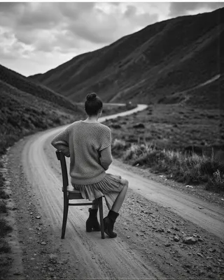 Black and white photo of a woman sitting on a wooden chair on a dirt road, facing away, wearing a knitted sweater and pleated skirt with a winding road and hills in the background.