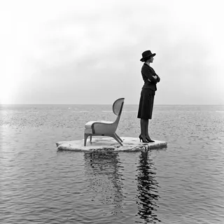 Black and white photograph showing a woman in formal attire and a wide-brimmed hat standing on a floating platform with a modern chair beside her, reflected in calm water under a foggy sky.