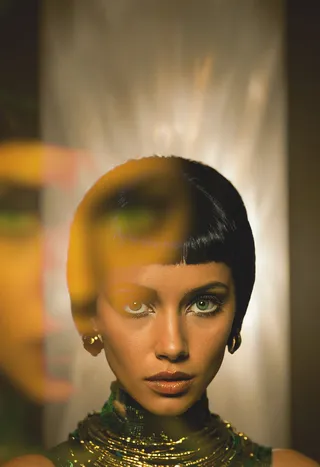 Portrait of a woman inspired by Cleopatra with short sleek black hair, piercing emerald-green eyes, golden-brown skin softly illuminated by warm chiaroscuro lighting, wearing gold-accented jewelry and garment.