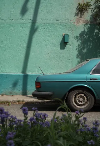 Rear side view of a turquoise 1980s car parked on a street with a textured green building wall and violet flowers in foreground under side lighting and long shadows